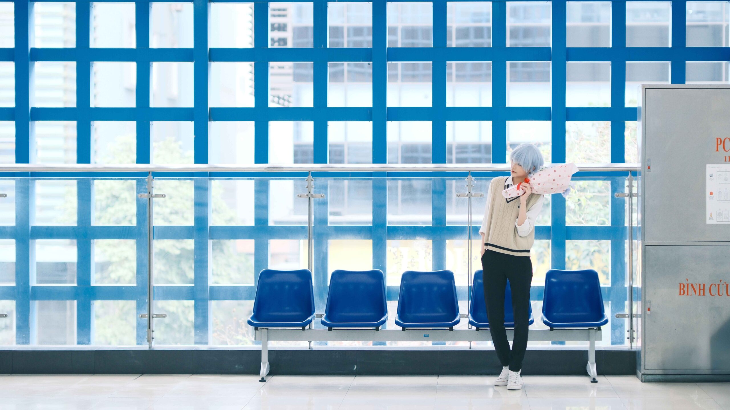 A stylish individual with blue hair stands by blue chairs indoors, holding a pink umbrella.